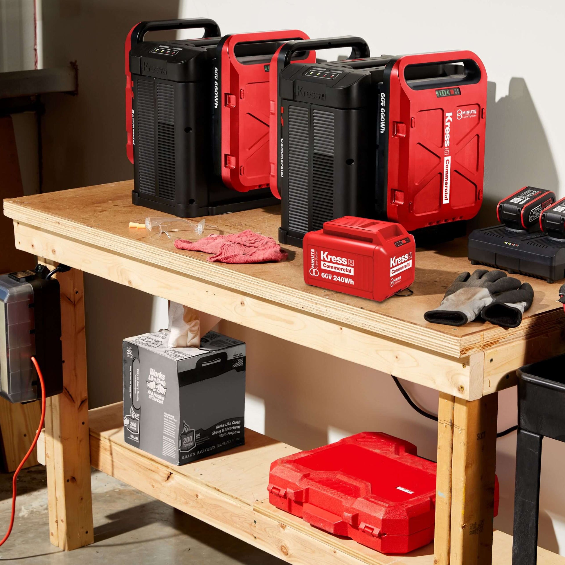A set of Kress 60v cordless tools, including two red and black portable power stacks, two battery packs, a red battery charger, a red tool case, and a red work glove, arranged on a wooden workbench in a workshop setting.